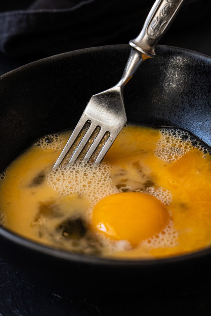 Close up of raw liquid eggs in a black bowl being mixed with a fork